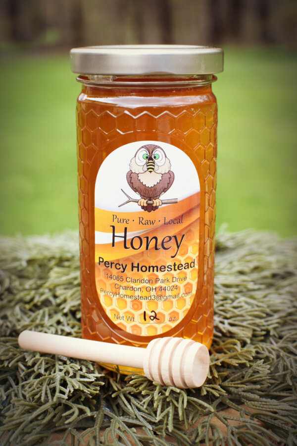 12 Ounce Jar of Honey with a Hexagon pattern on the jar, with a Percy Homestead product label. A dip stick is in front of the jar and the jar is sitting on a bed of pine needles with a green background.