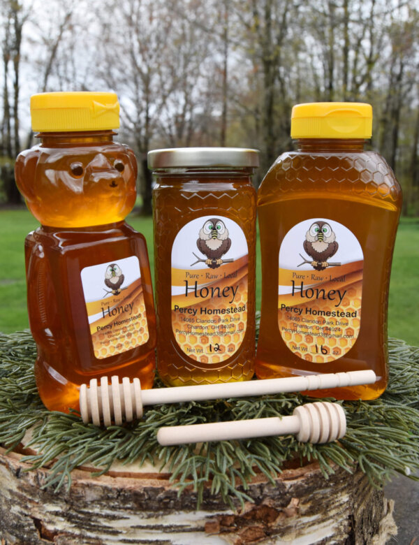 Three jars of honey on a stump with green pine needles, 1 bear bottle, one hourglass shaped bottle and on glass cylinder jar all have labels reading Percy Homestead. One large honey dipper and one small dipper are in front of the bottles.