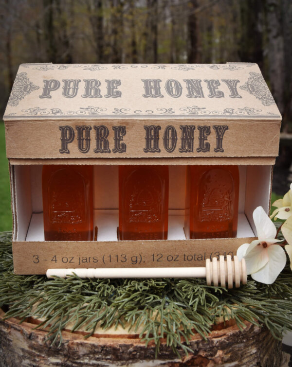 4 oz gift box with 2 muth bottles on pine needle base and treed background, white flower by the box and a 6" dipper stick in front of the box.