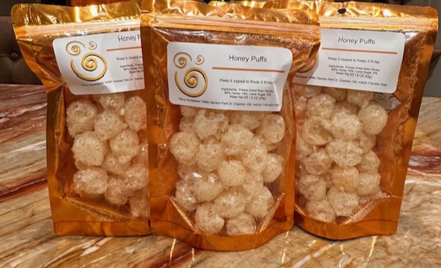 A clear plastic, resealable stand-up pouch filled with small, round, puffed honey treats. The label on the front reads “Honey Puffs” and includes a simple spiral bee logo, ingredient list (freeze-dried raw honey, cane sugar, water), and a local Chardon, Ohio address. The bag is displayed alongside similar packages on a wooden shelf.