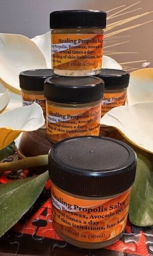 A small stack of amber glass jars labeled “Healing Propolis Salve” is arranged in a pyramid display. The jars have black lids and warm-toned labels listing ingredients like propolis, beeswax, and avocado oil. They are surrounded by soft white flower petals and green leaves, creating a natural, soothing aesthetic. The lighting is warm and highlights the golden color of the salve through the glass.